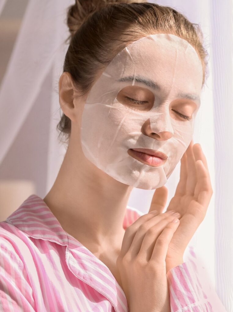 woman in pink suit wearing a face sheet mask with her eyes closed